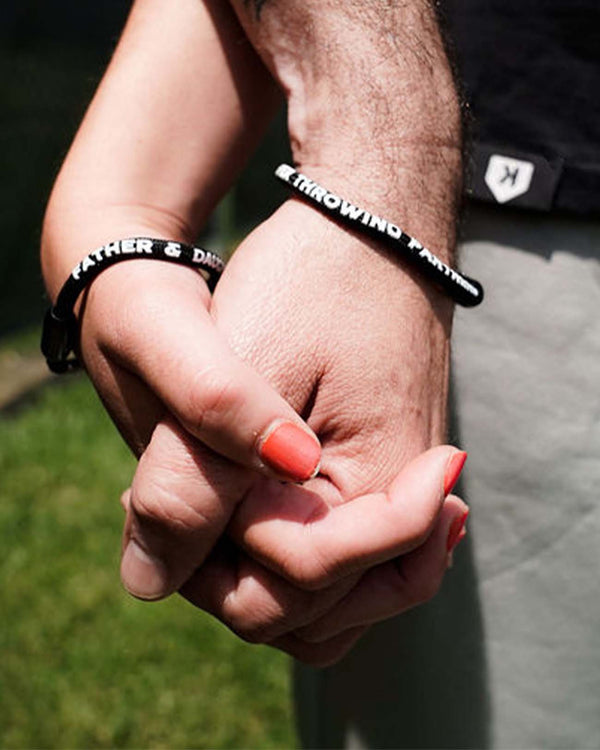 Father and daughter holding hands, both wearing black cord Pitchers Only bracelets reading FATHER & DAUGHTER: FOREVER THROWING PARTNERS