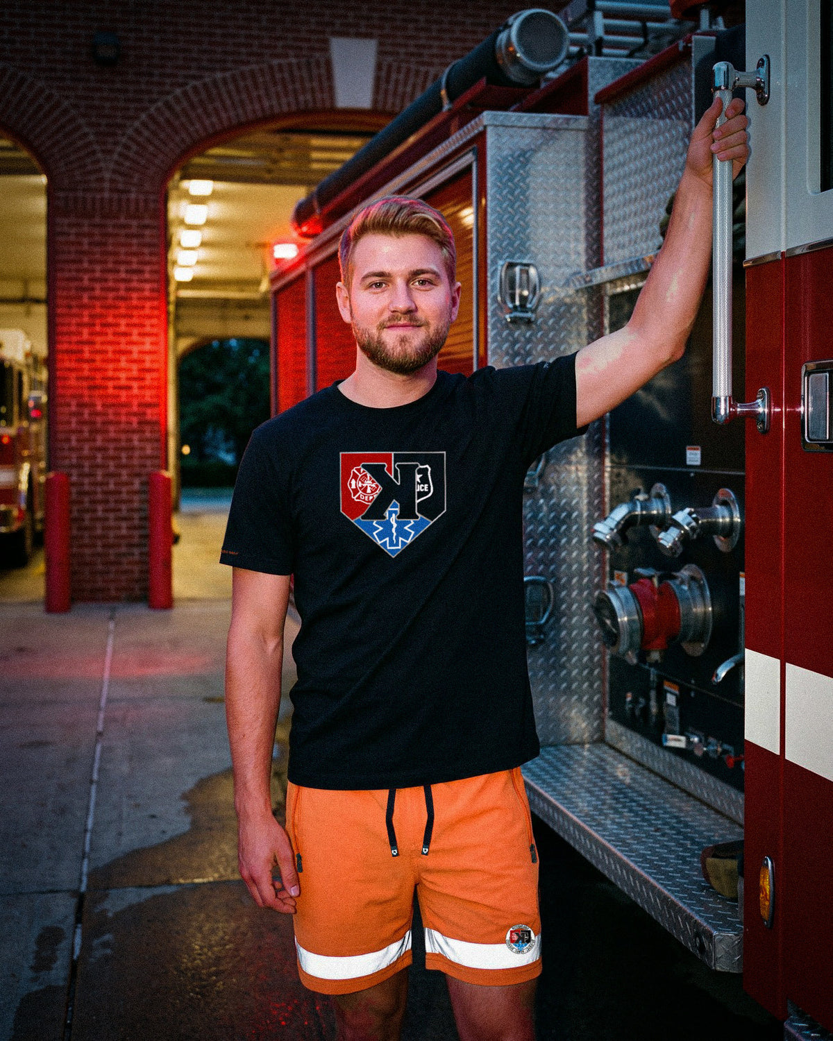 Model in the First Responders Tee and Safety Orange Shorts holding the grab rail of a fire engine - Pitchers Only